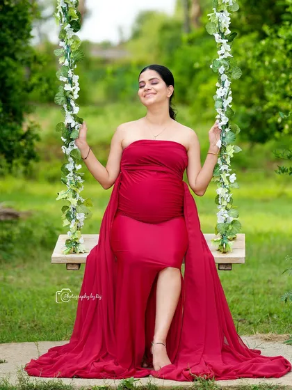 A serene and joyful portrait of a mother-to-be in a flowing red gown, seated on our floral swing. This setup in our private garden is perfect for capturing relaxed, candid moments that radiate happiness.
