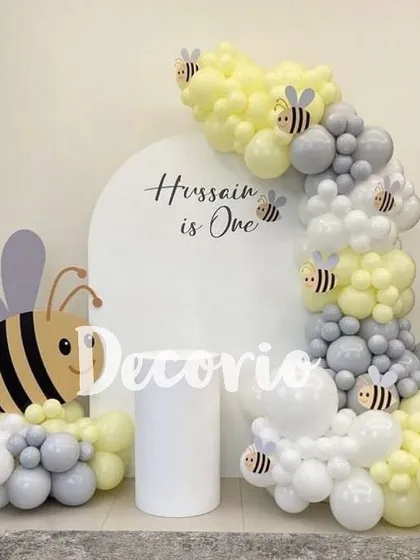 A cute "Hussain is One" bee-themed first birthday setup. The decoration includes a large bee cutout and a balloon garland in yellow, grey, and white, resembling a honeycomb.