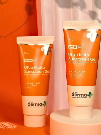 Two tubes of The Derma Co sunscreen are elegantly staged on pedestals against a warm, draped background, creating a sophisticated product display.