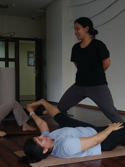 An instructor guides a student through a supine hip opening stretch. These immersive practices focus on releasing deep seated tension in the hips for greater mobility.