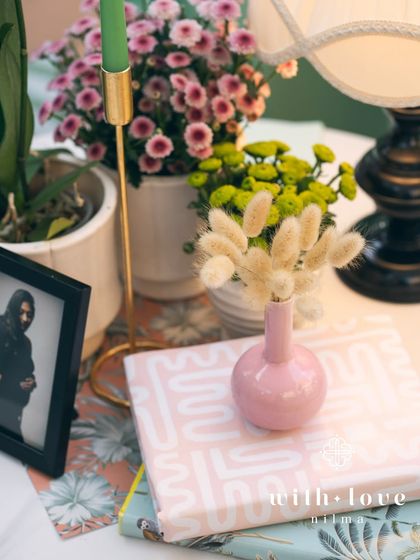 A close-up of a tabletop detail from 'The Conservatory' baby shower, including a small pink vase, a framed photo, and colorful flowers.