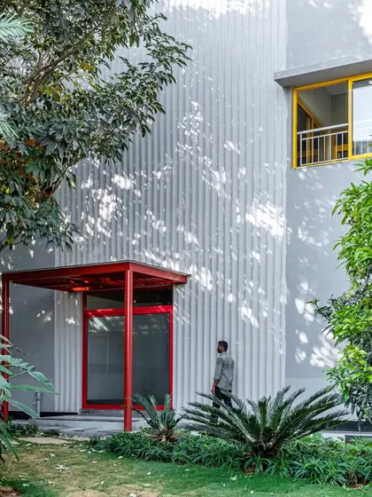 The entrance to L'Atelier is marked by a simple red portal, set against a corrugated wall texture. The building is nestled among existing trees, preserving the site's natural character.