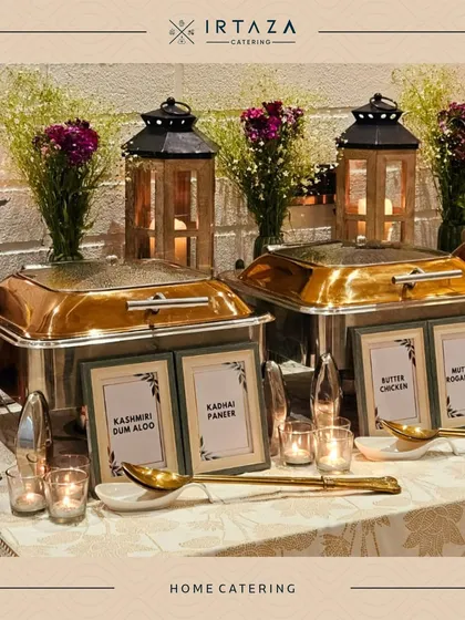A classic home catering arrangement with silver chafing dishes labeled with "Kashmiri Dum Aloo," "Kadhai Paneer," and more. The wooden lanterns and baby's breath flowers add a rustic yet elegant touch.