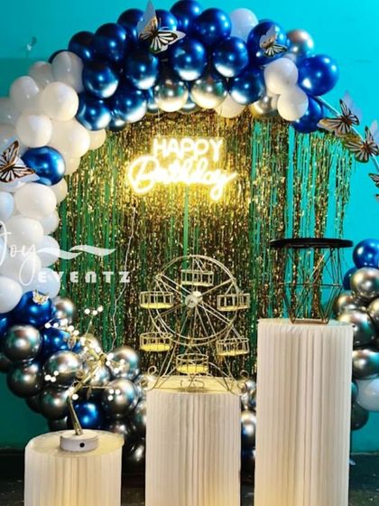 This stylish balloon ring decoration uses a combination of metallic blue, silver, and white balloons. The backdrop is a silver foil curtain, and the setup includes fluted plinths and a decorative Ferris wheel prop.
