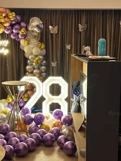 A 28th birthday party in a living room. This decoration includes a circular balloon arch in purple, gold, and silver, a neon sign, and marquee numbers.