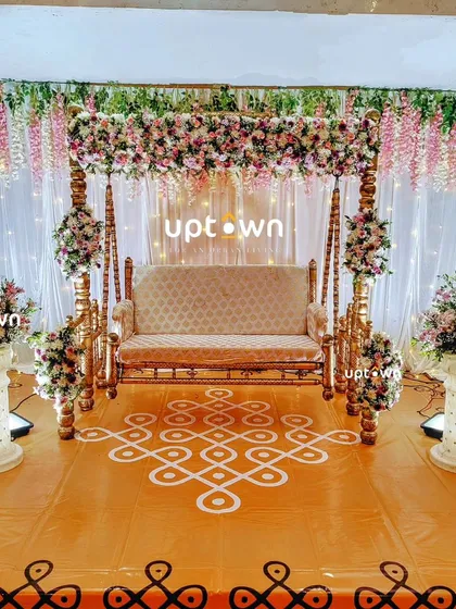 I specialize in traditional baby shower decorations that feel both classic and beautiful. This setup features an ornate wooden swing decorated with hanging flowers, set against a backdrop of white drapes and a traditional kolam floor design.