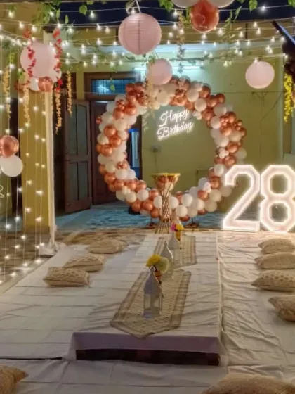 An intimate terrace party setup for a 28th birthday. The decoration includes a rose gold and white balloon ring, a neon sign, and a canopy of fairy lights over a low seating arrangement.
