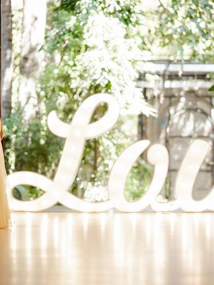 A cinematic portrait of a couple standing by a large, illuminated "Love" sign. The soft lighting and elegant pose make for a dramatic and romantic photograph.