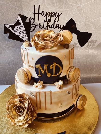 A sophisticated two-tiered cake for a man's birthday. It features a black and gold theme with tuxedo toppers, gold drip, and gold-dusted macarons, perfect for a formal celebration.