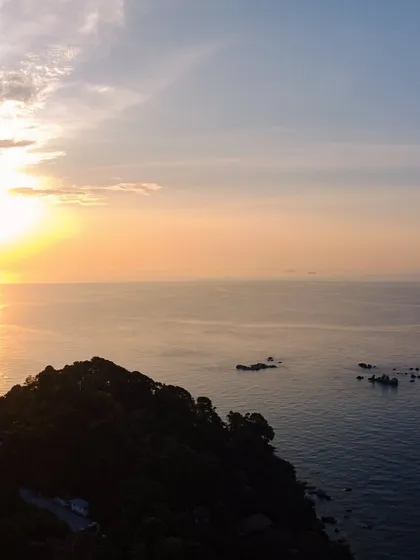 An aerial view of the sun setting over the ocean off the coast of Bintan Island, with the silhouette of a hill in the foreground.