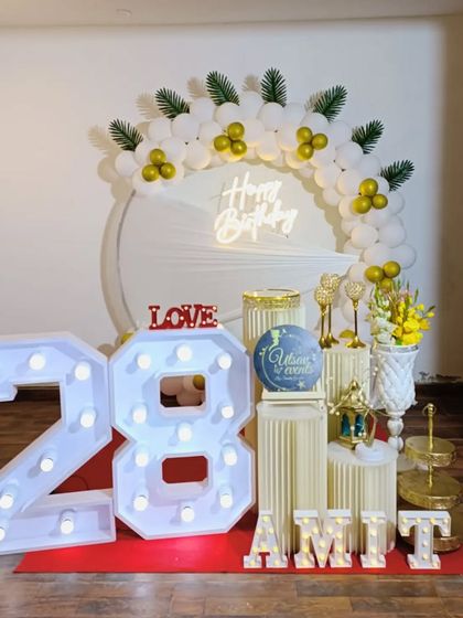 A beautiful white and gold themed birthday decoration. The setup includes a balloon arch with tropical leaves, a neon sign, and large marquee numbers for the age.