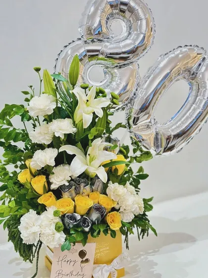 A birthday flower box arrangement with yellow and white flowers, silver-sprayed roses, and number balloons. This is a great example of a personalized birthday centerpiece.