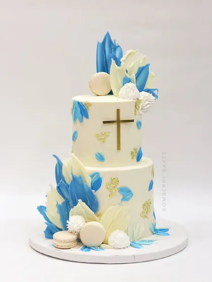 A two-tier baptism cake in white and blue, decorated with abstract chocolate sails, macarons, and a simple gold cross.
