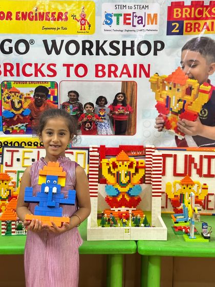 A smiling girl displays her blue and orange LEGO Ganesha in front of a table filled with other amazing creations from the workshop.