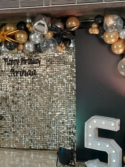 A glitzy birthday setup featuring a silver sequin wall, a balloon arch in black, gold, and silver, and a large light-up number '5'.