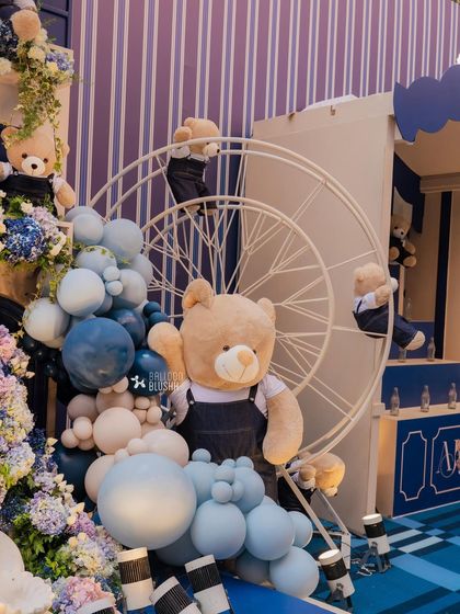 Teddy bears climb a Ferris wheel next to a cascade of blue and white balloons. This playful detail adds a dynamic element to the overall birthday party decor.