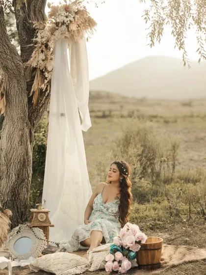 A dreamy, boho-inspired solo portrait. The mother-to-be sits in a beautifully decorated outdoor setting, with dried flowers, drapes, and a mountain backdrop creating a whimsical and romantic scene.