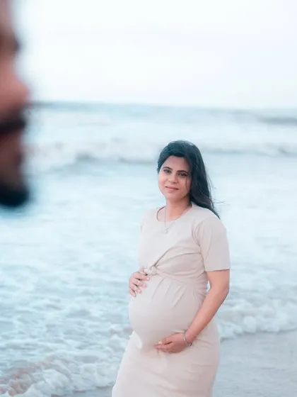 A beautiful solo portrait of the mother-to-be by the sea. The focus is entirely on her, her gentle smile, and the baby bump she lovingly holds, with her partner softly blurred in the foreground.