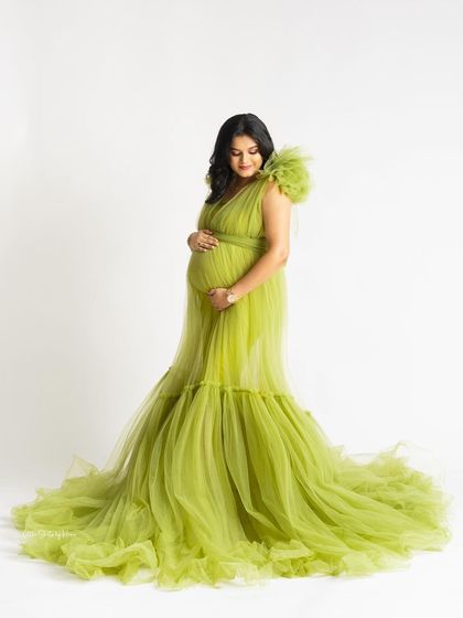 A full-length view of a gorgeous lime green tulle gown with a mermaid-style train. The mother-to-be looks down at her bump with a serene expression against a clean white background.