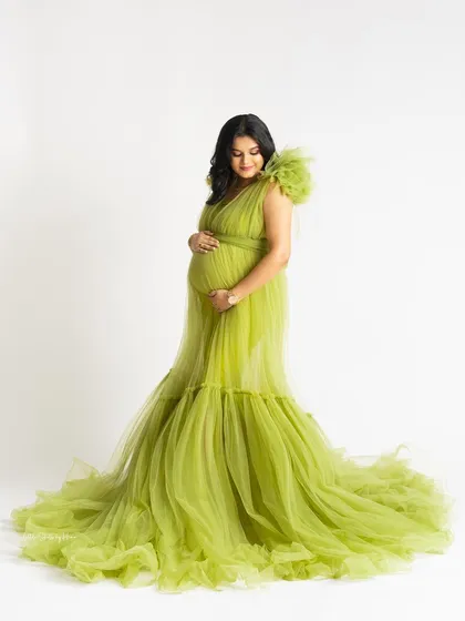 A full-length view of a gorgeous lime green tulle gown with a mermaid-style train. The mother-to-be looks down at her bump with a serene expression against a clean white background.