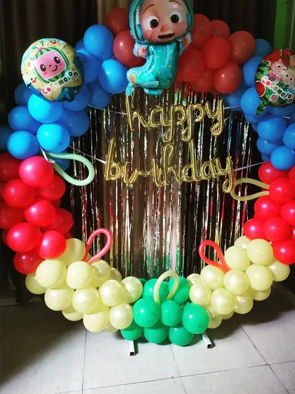 A Cocomelon-themed birthday decoration with a circular balloon frame. The colorful setup uses red, blue, yellow, and green balloons with character foil balloons.