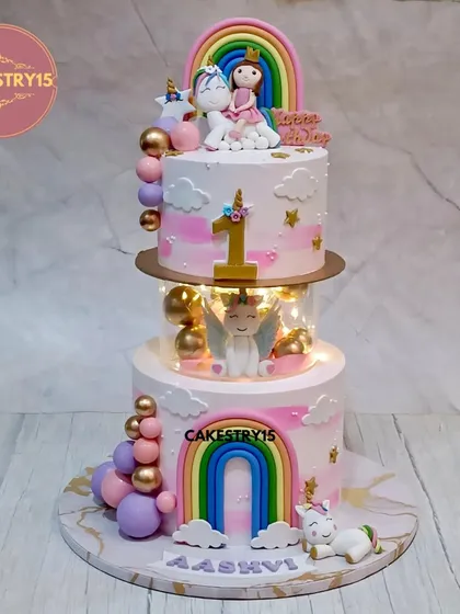 A magical first birthday cake featuring a two-tier unicorn and rainbow theme. The centerpiece is a glowing LED spacer with a winged unicorn, complemented by a topper of a girl riding a unicorn, fondant rainbows, and fluffy clouds.