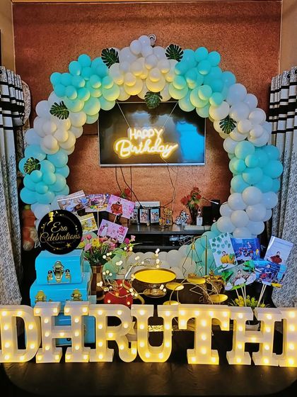 A first birthday home decoration with a circular balloon arch in pastel blue and white. The setup includes a neon sign and personalized photo displays.