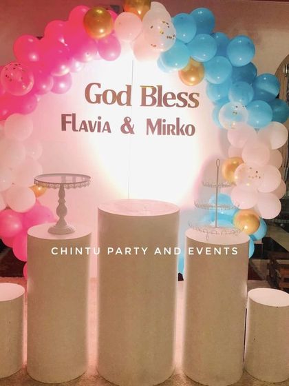A simple and clean backdrop for a First Holy Communion. The setup uses a backlit circular panel with personalized names and a pink, blue, and white balloon arch.