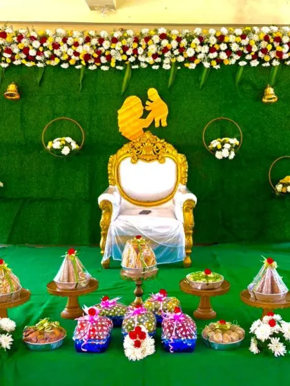 A full view of a baby shower stage, featuring a lush green backdrop, a central throne, and a beautiful arrangement of traditional sweets and offerings in the foreground.