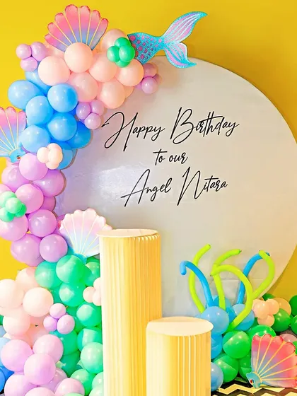 A pastel mermaid theme for Angel Nitara's birthday. This setup features a clean white backdrop with custom text, surrounded by a beautiful balloon arrangement with mermaid tail and shell accents.