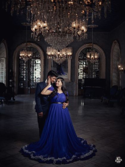 A moody and dramatic pre-wedding portrait in a grand hall. The deep blue gown with a ruffled trail looks stunning under the dim light of the chandeliers.