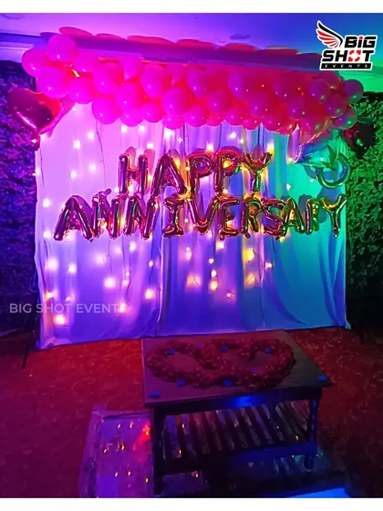 A classic anniversary celebration setup in a party hall, featuring a backdrop of white curtains with warm fairy lights, a bold 'Happy Anniversary' foil balloon, and a red balloon arch.