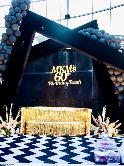 The modern stage for the 60th birthday bash, featuring a geometric black frame, balloon garlands, and a gold fringe sofa.
