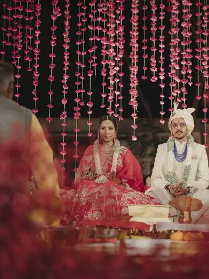 The couple seated for their wedding ceremony under a canopy of cascading pink floral strings. This intimate shot captures a serene moment amidst the beautifully detailed and textured decor.