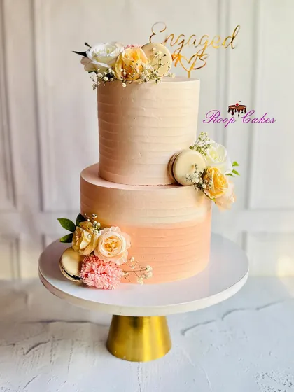 A sweet beginning to forever. This two-tier engagement cake features a warm peach ombre effect, adorned with fresh flowers and macarons for a touch of modern elegance.