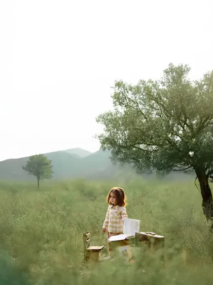 A wider view of the little girl with her miniature desk in the misty field. The composition emphasizes the solitude and wonder of a child lost in her own imaginative world, surrounded by the quiet beauty of nature.