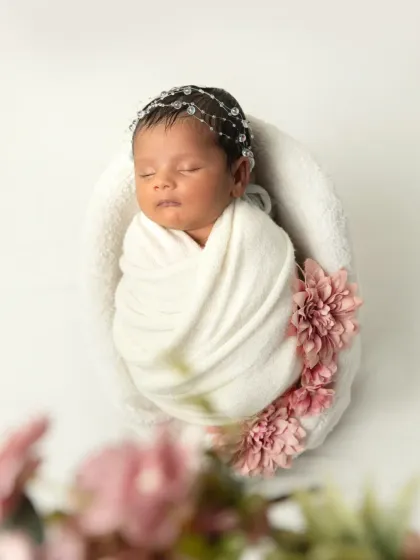 An angelic portrait of a sleeping newborn swaddled in white, with a delicate crystal headband. The blurred flowers in the foreground add a soft, dreamy feel.