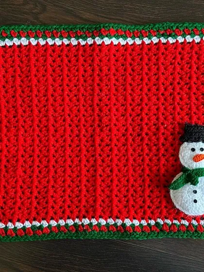 A bright red rectangular placemat with a snowman appliqué. The red, green, and white border adds an extra festive touch.
