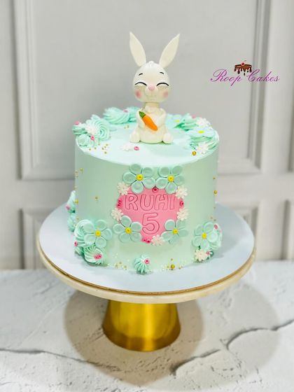 A sweet bunny-themed cake for a 5th birthday. This pastel green cake is decorated with flowers and topped with an adorable, handcrafted fondant bunny holding a carrot.