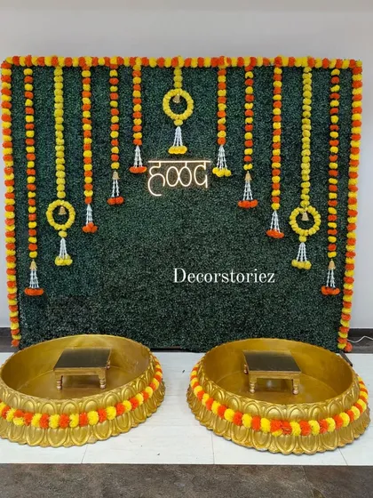 We designed this complete Haldi ceremony setup featuring a lush artificial grass wall backdrop, traditional marigold garlands, and a bright neon 'हळद' sign. The two large golden Urlis and matching patlas (stools) are also available from us on a rental basis.