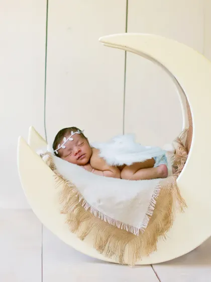 A classic and serene newborn portrait. The baby is dressed as a little angel, sleeping on a large, white crescent moon prop.