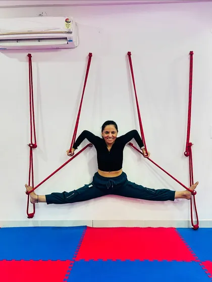 A smiling student enjoys the deep stretch of a supported wide-legged split using the wall ropes. This method helps to safely increase hip flexibility.