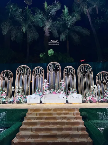 This magnificent outdoor stage is designed for a grand statement, with towering gold birdcage-inspired structures, lush pastel florals, and a dramatic staircase entrance.