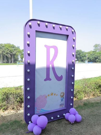 A purple marquee sign with the initial 'R' stands as a personalized decor piece at an outdoor event, complemented by a small cluster of purple balloons.
