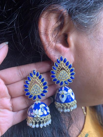 A grand pair of Meenakari jhumkas in royal blue. The stud is shaped like a peacock feather and is adorned with stones, while the dome has beautiful white floral painting.