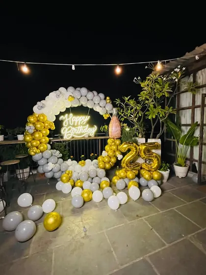 This 25th birthday was celebrated on a terrace with a custom balloon arch in gold, silver, and white. The decor features a 'Happy Birthday' neon sign and large '25' foil balloons to highlight the milestone.