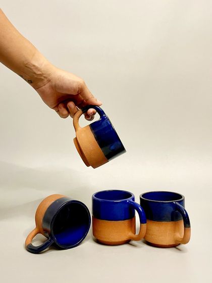 A hand holds one of my cobalt blue and terracotta mugs, with others arranged below. These durable, handmade mugs are perfect for your daily coffee or tea.