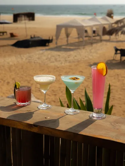 A lineup of colourful cocktails on a wooden railing overlooking the beach. This shot is all about the location, selling the experience of sipping drinks with an ocean view.