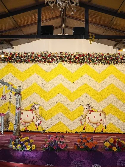 A large-scale floral backdrop for a Sreemantham with a yellow and white chevron pattern made of fresh flowers. The stage includes a decorated cradle and cow motifs for a traditional touch.