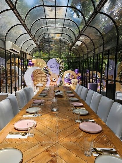 A long shot of the glasshouse venue, showing the dining table setup in front of the beautifully decorated baby shower stage.